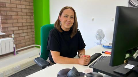 Kenieta Domaille pictured sat at her desk, smiling at the camera in a black top. She has shoulder length brunette hair with one side tucked behind her ear.