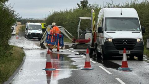 Workers in orange uniforms and white helmets are attending the water burst. Two company vehicles are parked on the side of the road. Three orange and white cones have been put around the burst. One of the workers is carrying blue road barriers.