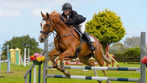 Justin Ogier and his brown horse jump over a grey, white and green fence during a showjumping competition. Justin is wearing a black jacket, white jodhpurs and black riding boots. He has a black helmet on. Other showjumping fences are dotted around the tree-lined field.