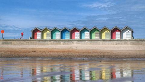 Blyth beach at low tide, looking back to the beach from the water. Colourful huts line the beach and a man with a red top runs along the promenade. The huts are reflected in the water and the sky is very blue.