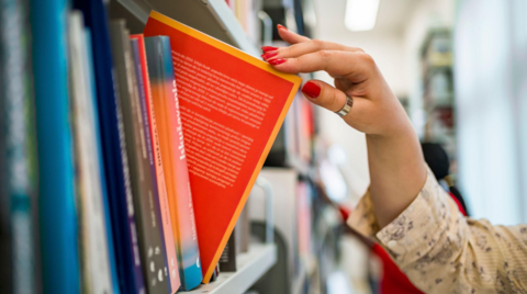 A shelf of books is on the left of the image and a woman has fingers on one mainly red book that she has partially pulled from the shelf.