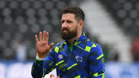 Josh Vickers is pictured waving with a straight face. He is in a blue and green Derby training top. 
