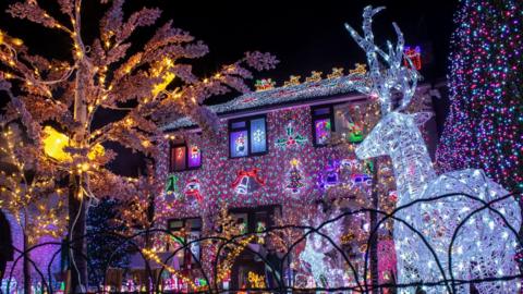 A house covered in bright coloured blue and purple lights. To the right there is a bright white shining reindeer illuminated by wires. The windows have snowmen and candy cane lights, as well as other Christmas objects.