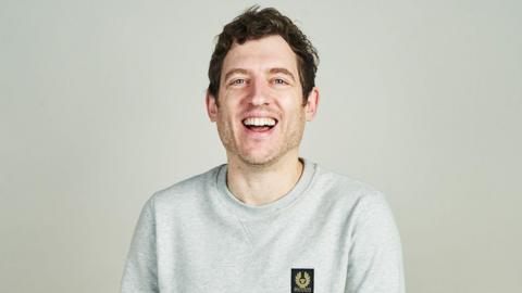 Elis James headshot. He is wearing a grey sweatshirt and laughing.