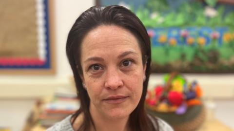 A woman with dark hair and brown eyes not smiling and looking directly at the camera in a room with out of focus toys in the background