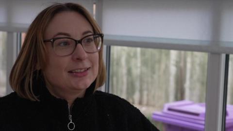 Heather Ainsworth being interviewed in her grandmother's home. She has shoulder length brown hair and is wearing glasses and a black top.