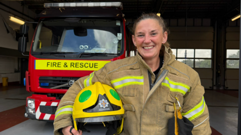 Tracey Laws, an on-call firefighter, pictured in her uniform. It consists of a beige-coloured, heavy-duty jacket with h-vis stripes, and a yellow helmet which bears the fire service's logo. She is smiling and standing in front of a fire engine, which has Fire and Rescue emblazoned in capital letters on a yellow background.