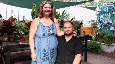 A woman in a blue floral dress and headscarf stands beside a man seated in a wheelchair, wearing a black shirt. Both are inside a greenhouse filled with potted plants.