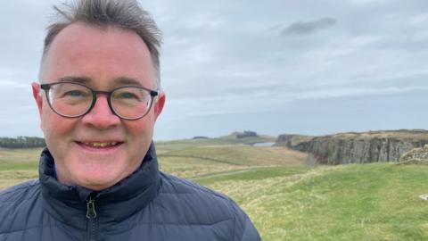 Tony Gates stands with a stretch of Hadrian's Wall rising on a crag behind him. He is a man in his 60s with glasses and short hair smiling and with a blue jacket zipped up to his neck.
