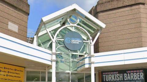 Main sign of Salford Shopping Centre over its entrance. It is part of a white frame in front of a glass panel.