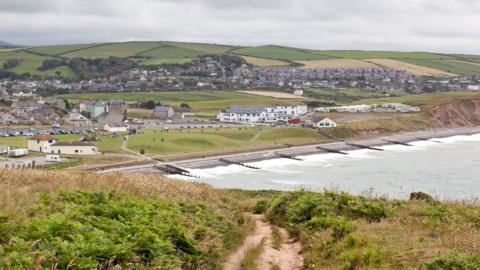 St Bees beach is pictured in the background with groups of houses near the water's edge with groynes lining the beach itself, and in the foreground is a coastal path surrounded by vegetation, on a cloudy day.