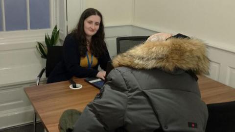 Two women sit on either side of a brown-topped desk, in a small room with cream walls in a small room, that has a window on the left. The woman in the foreground has her back to the camera and is wearing a parka-style coat, as she has a conversation with the other woman, who is wearing a security pass.