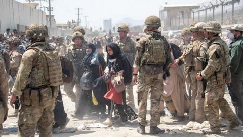 British armed forces work with the US military to evacuate eligible civilians and their families out of Afghanistan on August 21, 2021. Soldiers on dusty road between wire fences topped with razor wire escorting two women dressed in black