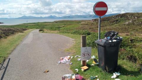 A wheelie bin overflowing with litter, next to a "No Entry" sign. It sits next to single track road which leads off into the distance. In the background is a stretch of water and a picturesque, mountainous landscape. 