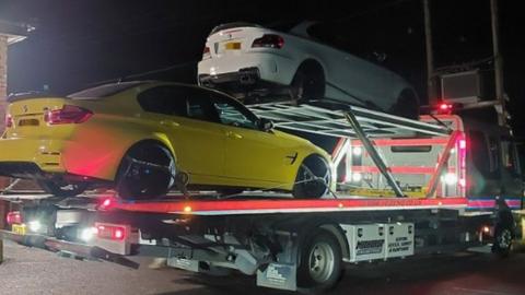 Flatbed truck with yellow car on the bottom and white car on a ramp above