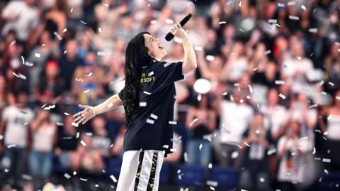 Billie Eilish - a young woman with long, dark hair, wearing a dark t-shirt and white jogging trousers, is seen side view, singing into a microphone in front of a large audience, with white confetti falling around her.
