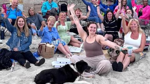 Large group of people seated and standing on a sandy beach in front of a concrete wall, raising their arms toward the camera. A dog lies on the sand in the foreground, with picnic items and bags scattered around.