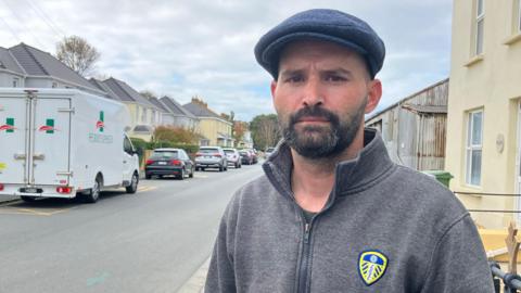 A man in a grey zip up hoodie. He is looking at the camera. He is wearing a blue cap and has a black beard. Behind him is a road. There are cars parked along it. The sky is grey. 