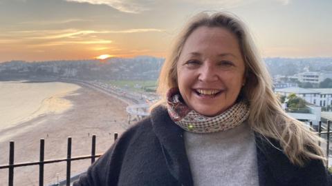 Katya Maddison wearing a grey sweater, a scarf and a black coat, with Torquay and Paignton seafront in the background. The sun is setting over Paignton and the sky is glowing orange and yellow