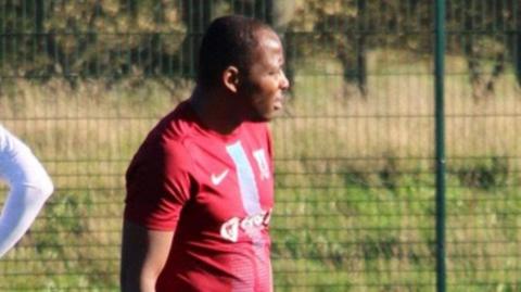 A man wearing a red football top stands on a field looking away from the camera.