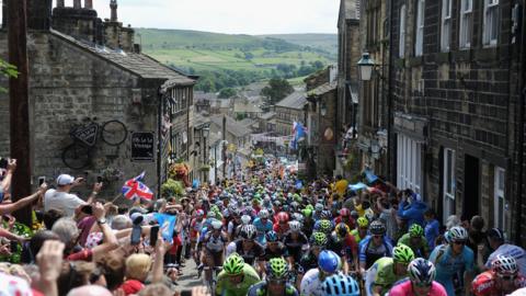 A steep hill with bricked buildings on either side. The street between the buildings is filled with professional cyclists on their bikes. They are wearing an array of colourful helmets. Stood at the side are crowds of people. One person is waving a union jack flag and others are taking pictures.
