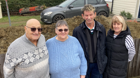 Brian and Jane Goodger are standing on the left next to their neighbours Roger and Sandra Wright. Behind them is a mount of dirt and a driveway with a car parked on it.