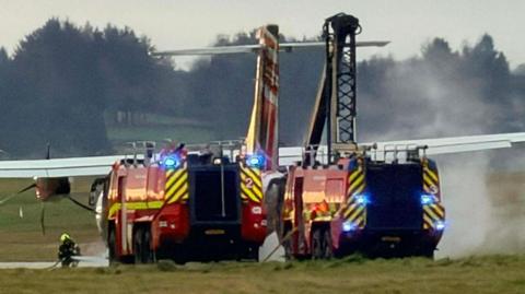 Two airport fire trucks with flashing blue lights positioned behind a turboprop aircraft on a grassy area, with smoke or steam rising near the plane and a firefighter working on the ground