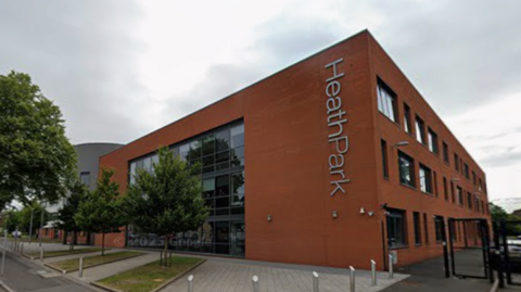 A brick three-storey building, with large windows on one side which almost cover its entire height from the floor to the roof. There are metal posts surrounding a paved area which also has areas strips of turf and trees spaced evenly in front of the building.