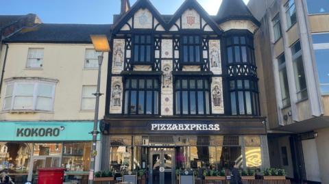 A historic building with Pizza Express signboard - with frontage features five painted statues of historical figures, including: Aethelwold Bishop of Winchester, King Peada of Mercia, Henry VIII, Prince Rupert and the Earl of Essex.