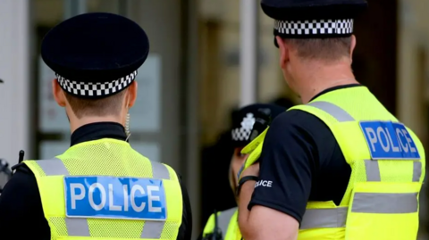 Two police officers wearing high visibility yellow vests with the word police written on the back. They have the backs to the camera and are also wearing black police caps.