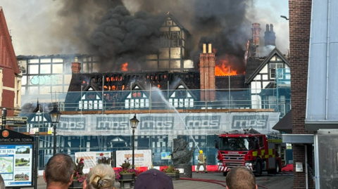 An old white building with black timber frames surrounded by scaffolding with flames and smoke coming from the roof