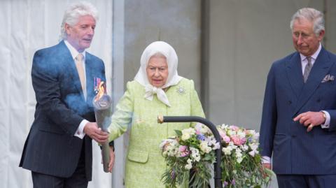 Man in dark suit and thick white hair hands a flaming torch to the late Queen Elizabeth who is wearing a white headscarf and lime green coat to ignite the first beacon during celebrations to mark her Diamond Jubilee. A large bouquet of pink, white and lilac flowers are in front of her. Prince Charles, wearing a navy, double-breasted suit, stands next to her, and has a bemused smile on his face.