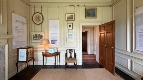 A room with white walls, a big oak door and various paintings on the wall.