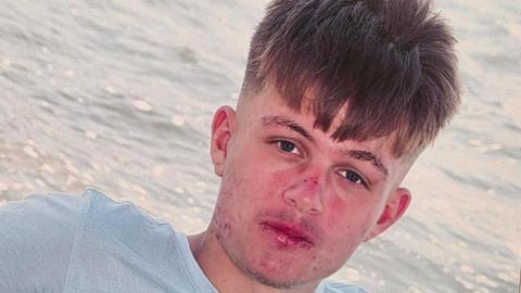 Joshua Ingram, a 16-year-old boy, is wearing a t-shirt and sitting in front of the sea, with waves visible behind him.