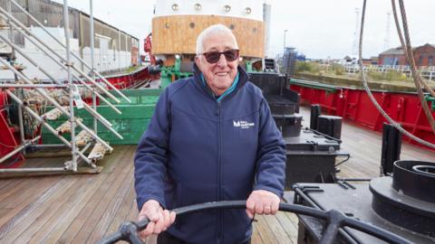 An man with short white hair and sunglasses holds onto a black metal bar on the deck of a boat.