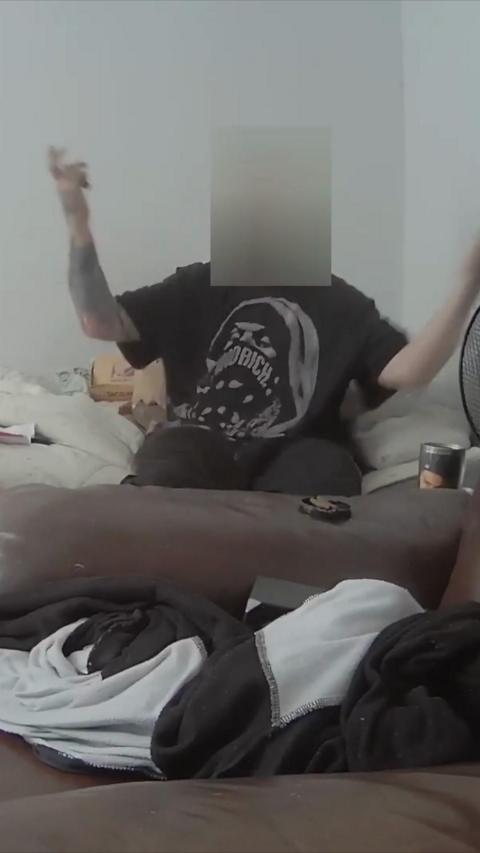 A man in a dark blue t-shirt sits on his bed with his face blurred. 