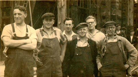 A black and white photograph of six workers at the Goole Shipyard. The men are wearing overalls and are smiling at the camera. Three of them are wearing flat caps and one is smoking a pipe.