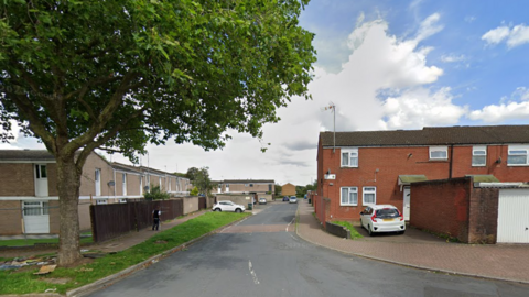 A residential road with white lines down the middle has two storey red and grey brick houses either side and a large tree on the left