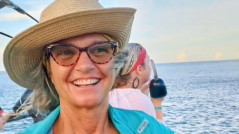 A photo of Lynette Hooker smiling while on a body of water. She is wearing a turquoise-like coloured shirt, glasses, and a woven straw hat.