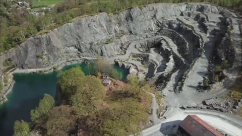 An aerial view of Elterwater Quarry. Layers of grey rock are to the right of a pool of water and some trees.