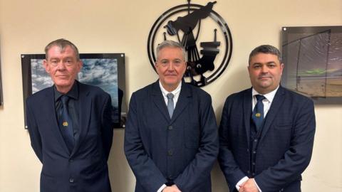 Three smartly dressed men in navy blue suits and ties are standing in front of the crest of Comhairle Nan Eilean Siar. It is a birlinn style medieval ship with a sail and oars.