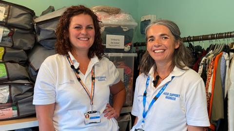 Two women wearing white tops and smiling at the camera. There are racks of clothes behind them. 