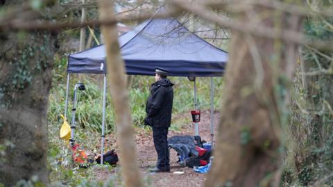 A police officer stands near a gazebo in a wooded area. The gazebo has lots of items under it on the ground.