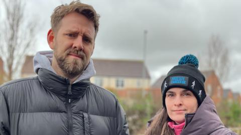 Chris and Nicola Day-Coiley stood together with winter coats on. Chris is wearing a black coat, has blond-ish hair and a beard. Nicola is shorter than him and wearing a black beanie with a long brown ponytail over one shoulder. She is wearing a grey/purple coat.