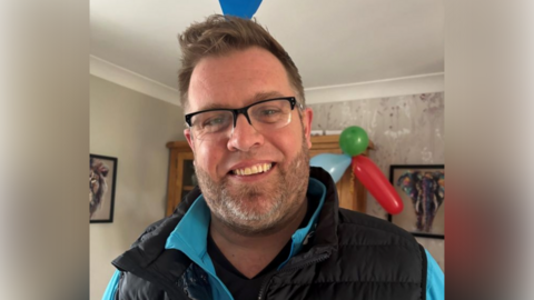 A head and shoulders photo of Zack Mitchell standing in a living room. He is smiling at the camera, wearing glasses, a black top, a blue zip-up jacket and a black, padded gilet. He has short brown hair and stubble on his face. 