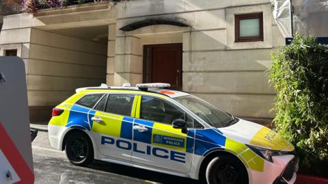 A yellow and blue police car is parked on a street in front of a doorway. The building it is parked next to is a beige yellow colour with a small window to the right next to a bush. 