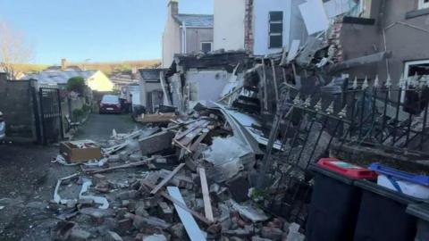 A lane behind houses is strewn with rubble, bins and blown-off metal black gates. There is a house with its walls blown off. 