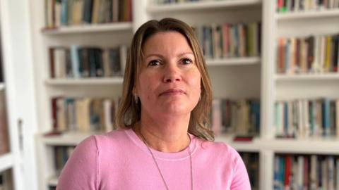A woman with shoulder length brown hair. She is facing the camera. She is wearing a pink top and a silver necklace. In the background are white shelves with books on. 