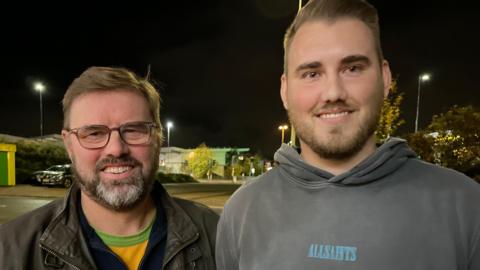 Two men standing side-by-side outside at night - the one on the left is wearing spectacles and a brown jacket with a yellow green t-shirt underneath. He has brown hair and a greying beard. The man on he right is about five inches taller, with brown hair and a beard. He is wearing a grey hoodie with ALLSAINTS written across the chest in light blue. The background is made up of street lights and trees.