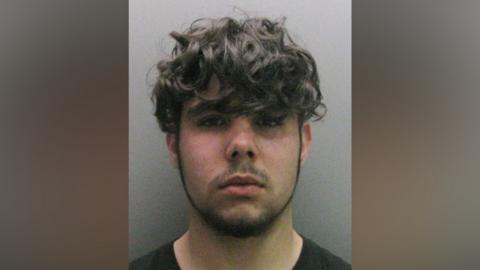 A mugshot of Dylan McNamara. He has brown wavey hair and stubble.